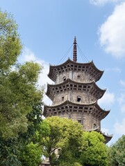 Naklejka premium Ancient stone pagoda piercing the sky under blue skies and white clouds, with lush green trees obscuring its base, evoking a timeless, historical ambiance
