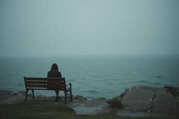 A solitary figure sits on a bench overlooking a misty ocean.