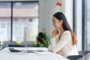 Asian businesswoman working at a desk, using a laptop while reviewing financial documents in a...