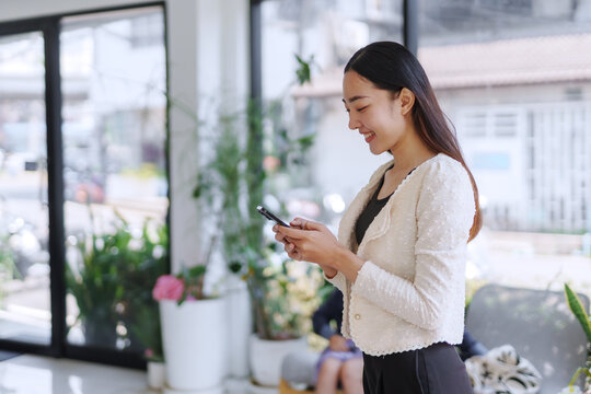 Smiling businesswoman using a smartphone while standing in a bright, modern office space filled with natural light and greenery - Powered by Adobe