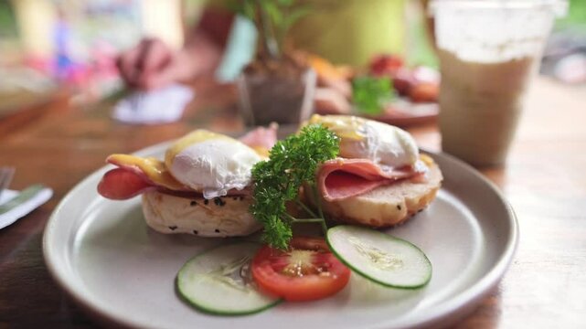 Plate of eggs benedict with poached eggs, ham, and hollandaise sauce on toasted English muffins served with sliced cucumber and tomato on wooden table in casual brunch setting