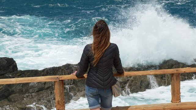 A woman with her back turned and long dark hair admires the waves crashing against the coast of the Ajui caves area on the island of Fuerteventura.