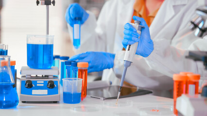 Fototapeta premium Two Asian female scientists wearing laboratory gloves sit at a table, carefully examining a test tube filled with blue liquid, using a microscope for detailed analysis in a research laboratory