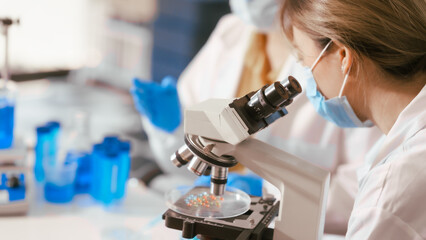 Two Asian female scientists wearing laboratory gloves sit at a table, carefully examining a test tube filled with blue liquid, using a microscope for detailed analysis in a research laboratory