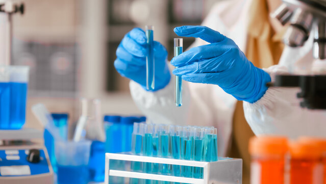 Two Asian female scientists wearing laboratory gloves sit at a table, carefully examining a test tube filled with blue liquid, using a microscope for detailed analysis in a research laboratory