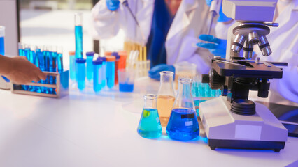 Two Asian female scientists wearing laboratory gloves sit at a table, carefully examining a test tube filled with blue liquid, using a microscope for detailed analysis in a research laboratory