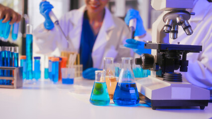Fototapeta premium Two Asian female scientists wearing laboratory gloves sit at a table, carefully examining a test tube filled with blue liquid, using a microscope for detailed analysis in a research laboratory