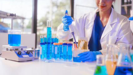 Fototapeta premium Two Asian female scientists wearing laboratory gloves sit at a table, carefully examining a test tube filled with blue liquid, using a microscope for detailed analysis in a research laboratory