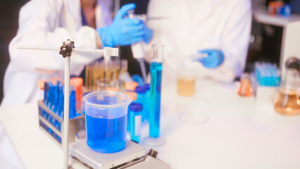 Fototapeta premium Two Asian female scientists wearing laboratory gloves sit at a table, carefully examining a test tube filled with blue liquid, using a microscope for detailed analysis in a research laboratory