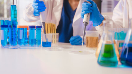 Two Asian female scientists wearing laboratory gloves sit at a table, carefully examining a test tube filled with blue liquid, using a microscope for detailed analysis in a research laboratory