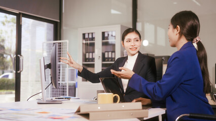 Two young Asian business woman sit at a table in the office, discussing marketing strategies, using a computer reviewing documents with charts and graphs, collaborating on new business plans at work