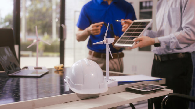 Two Asian businessmen wearing engineer hats table, discussing solar panels and wind turbines. They plan environmental projects, focusing on solar energy solutions and renewable energy installations