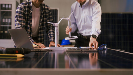 Two Asian businessmen wearing engineer hats table, discussing solar panels and wind turbines. They plan environmental projects, focusing on solar energy solutions and renewable energy installations