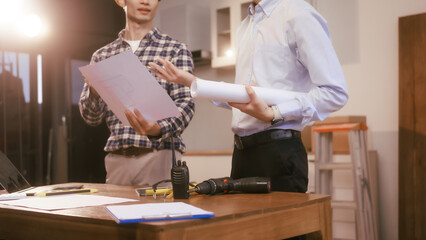 Two male engineers discuss a construction blueprint at a table in a modern office. They focus on...