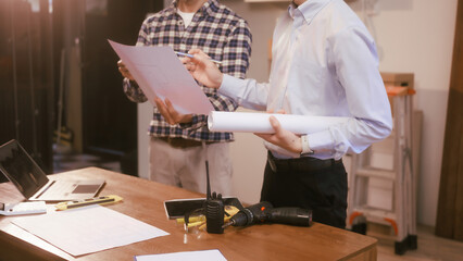 Two male engineers discuss a construction blueprint at a table in a modern office. They focus on...