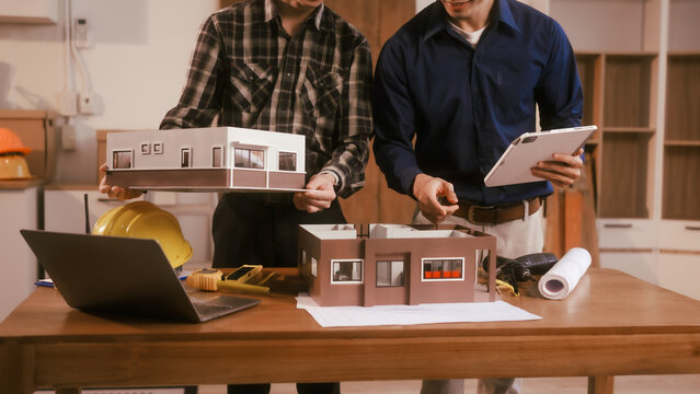 Two male engineers review blueprints in an office while discussing model house.They talk about construction projects, architectural design,roof structures,contractor coordination, building materials