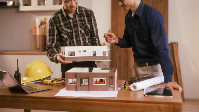 Two male engineers review blueprints in an office while discussing model house.They talk about construction projects, architectural design,roof structures,contractor coordination, building materials
