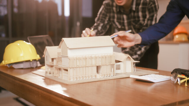 Two male engineers review blueprints in an office while discussing model house.They talk about construction projects, architectural design,roof structures,contractor coordination, building materials
