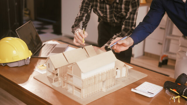 Two male engineers review blueprints in an office while discussing model house.They talk about construction projects, architectural design,roof structures,contractor coordination, building materials