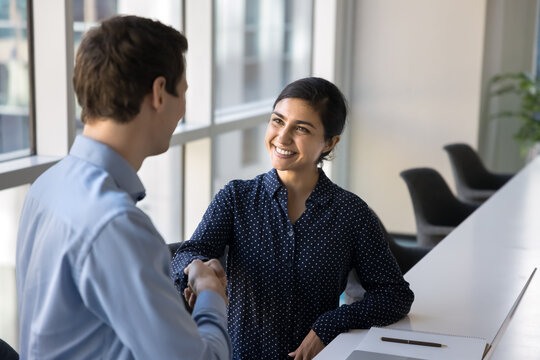 Indian and Caucasian professional teammates shaking hands sitting together at table in office. Cheerful female applicant passed successfully job interview, handshaking HR manager, accomplish meeting