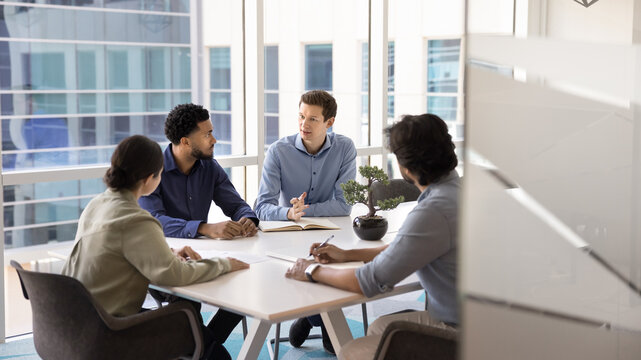Four diverse professionals talking, negotiating in contemporary meeting room, discussing deal, project details and updates, planning start-up launch, engaged in productive teamwork in corporate office