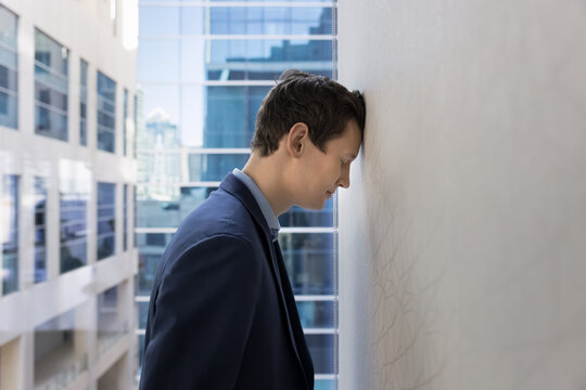 Young 30s businessman leaning against wall his head, feeling exhausted and overwhelmed, experiences emotional challenges faced in corporate world. Business failure, dismissal and professional burnout