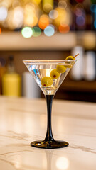 Martini with three green olives on a skewer sits on a marble counter. The background is a softly blurred, colorful bar setting