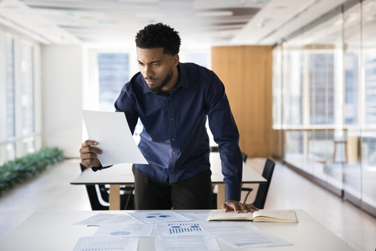 Focused African professional leans over table filled with charts, graphs, documents, reviewing report, engaged in strategic planning, data analysis and decision-making in contemporary coworking office