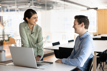 Two young multiethnic colleagues engaged in funny conversation in modern co-working office, discussing work-related tasks, joking, laughing, having friendly work relations, collaborating on project