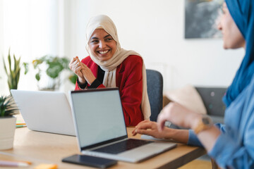Two women in hijabs are collaborating on a project. They're using laptops at a desk, brainstorming ideas and working together to achieve their goals.