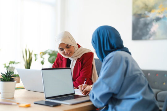 Two women in hijabs collaborate on a project. They're using laptops and taking notes, likely brainstorming or problem-solving together in a bright, modern office space.