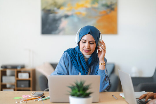 A woman in a blue hijab and headphones works on her laptop at a desk. She's focused and listening, likely attending a virtual meeting or working remotely from home.