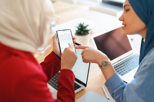 Two women in hijabs are collaborating on a project. They're using a phone to check their schedule and plan their next steps, ensuring they stay on track with their goals.