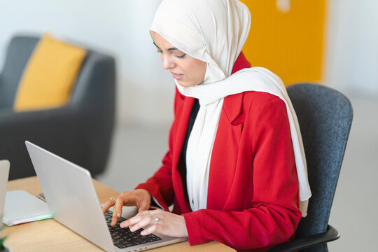 A woman in a hijab and red blazer is working on her laptop. She's focused on her work, likely completing a project or responding to emails.