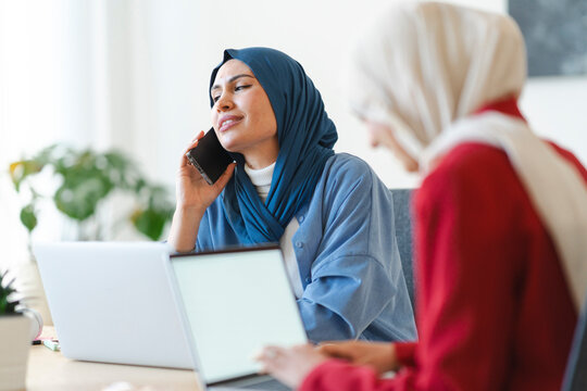 Two women in hijabs are working together. One is on a call, possibly discussing a project, while the other focuses on her laptop. They're collaborating to achieve a common goal.