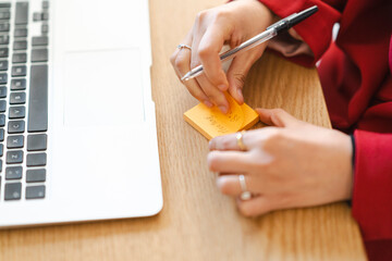 A woman is writing a reminder on a sticky note. She's using it to remember a meeting, so she doesn't forget while working on her laptop.