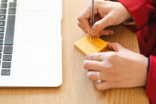 A person is writing "Meeting 18:00" on a sticky note. They're likely scheduling or reminding themselves of an upcoming meeting. A laptop sits nearby.