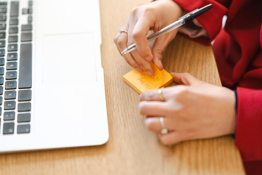 A woman is writing a reminder on a sticky note. She's using it to remember a meeting, so she doesn't forget while working on her laptop.