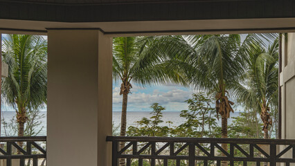 Ocean view from the resort's terrace. Spreading palm leaves against a blue sky and clouds. Turquoise sea. In the foreground are columns and latticed wooden railings. Malaysia. Borneo. Kota Kinabalu.