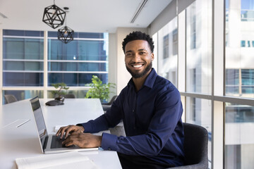 Fototapeta premium Portrait of successful professional African businessman, office employee sitting at table working on laptop in modern boardroom, look at camera, feel happy and proud with achievements, career growth