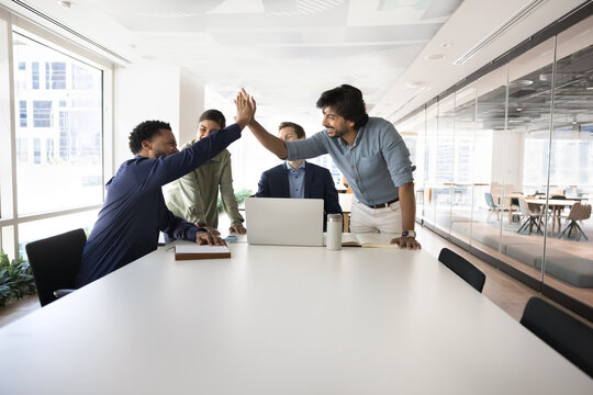 Happy multiethnic teammates gathered together in meeting room celebrate joint success, shared goal achievement with high five, gesture of appreciation, congratulate each other with professional growth