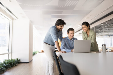 Three multinational colleagues gathered around laptop in modern coworking space, collaborate on...
