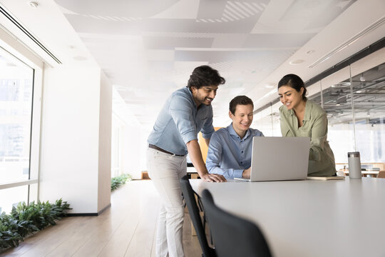 Three multinational colleagues gathered around laptop in modern coworking space, collaborate on project, discuss ideas, assign tasks and work on shared goal during briefing event in corporate office
