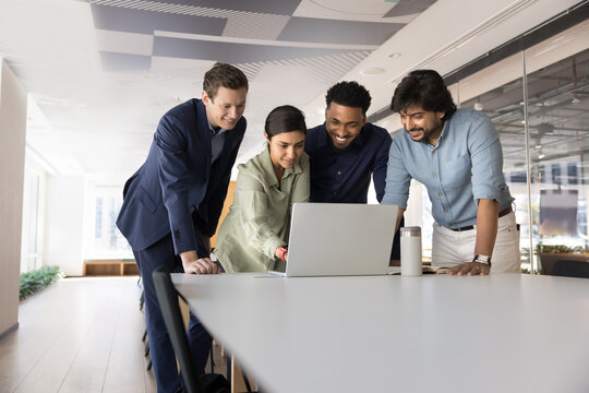 Four multinational businesspeople, teammates using laptop, tracking sales results, review new updates, working on joint on-line project gathered together in office. Teamwork using modern technology