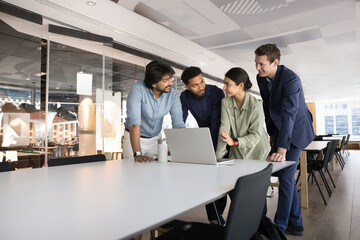 Four young multiethnic professionals gathered around laptop in corporate office, exchanging point, opinion or ideas, listening Indian female guiding, provides explanation regarding software or task