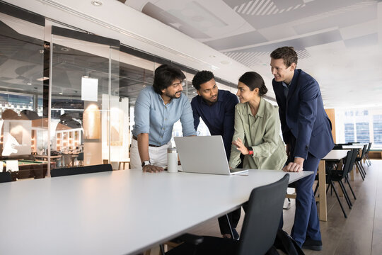 Four young multiethnic professionals gathered around laptop in corporate office, exchanging point, opinion or ideas, listening Indian female guiding, provides explanation regarding software or task