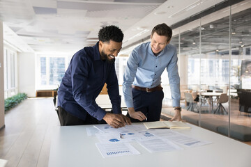 Two multiethnic professional teammates working together, analyzing business data, preparing financial report, reviewing charts for strategy development, smiling, discussing profit and statistical data