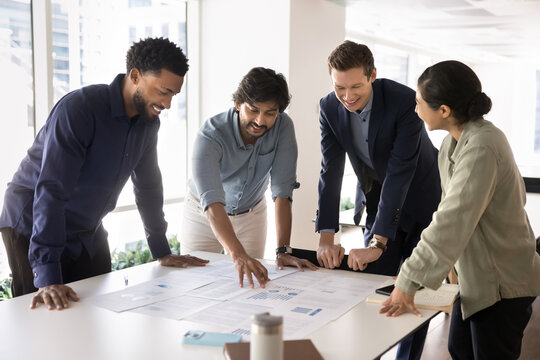 Multiethnic corporate team engaged in positive, productive meeting, gathered round desk with papers, analyzing business data or report, collaborating on project, reviewing performance metrics or plans
