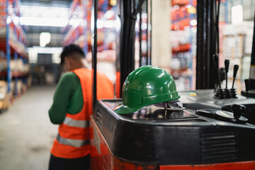 Forklift Operator in Warehouse with Green Hard Hat and Safety Vest in Background with Shelving...