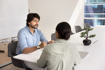 Obraz premium Two Indian teammates lead conversation about joint task completion or project updates seated at table in modern conference room. Middle-aged man bank advisor consulting female client during meeting